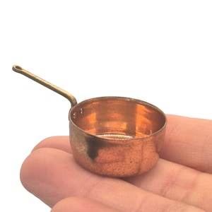 May include: A miniature copper saucepan with a brass handle. The small pot is round with a slightly flared rim. The handle is attached to the side and curves upwards. The pan is held in a person's hand.
