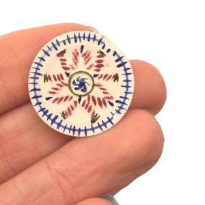Könnte beinhalten: Ein kleiner, runder, handbemalter Keramikteller mit floralem Design. Der Teller hat eine weiße Basis mit einem zentralen roten und grünen Blumenmuster, umgeben von blauen und grünen Akzenten. Der Teller wird in einer Hand gehalten.