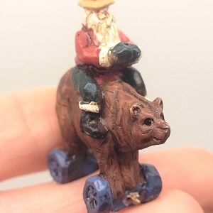 May include: A miniature figurine of a bear with a person riding on its back. The bear is brown with blue wheels. The person is wearing a red jacket, a yellow hat, and has a white beard.