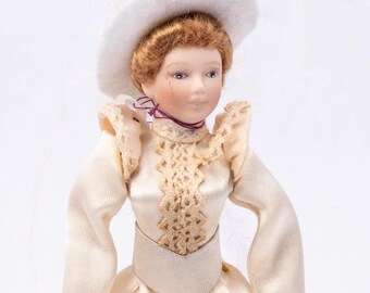 May include: A porcelain doll wearing a white dress with lace detailing and a white hat with floral accents. The doll has brown hair and painted blue eyes.