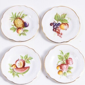 K&ouml;nnte beinhalten: Set aus vier wei&szlig;en Desserttellern aus Keramik mit goldener Verzierung. Jeder Teller zeigt ein anderes Fruchtdesign: Ananas, Trauben, Wassermelone und Erdbeeren.