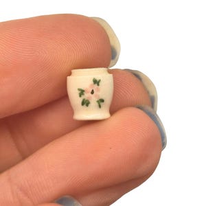 以下が含まれることがあります： ミニチュアのクリーム色のセラミック花瓶。ピンクの花と緑の葉のデザイン。小さな花瓶は、白い背景に対して2本の指の間に保持されています。
