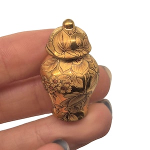 May include: A small, gold-colored decorative jar with a lid. The jar is adorned with a floral pattern in a darker gold hue. The jar is held between fingers, showcasing its size and intricate design. The jar is likely a miniature decorative item.