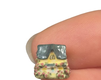 Może przedstawiać: Miniaturowy żółty domek z szarym dachem i różowymi kwiatowymi akcentami. Mały domek jest trzymany między palcami, prezentując jego mały rozmiar i szczegółowy design. Domek ma drzwi i okna.