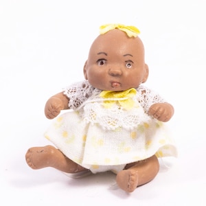 May include: A small porcelain doll with brown skin, wearing a white dress with yellow floral accents and a yellow bow. The doll has a yellow bow in its hair.