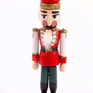 May include: A wooden nutcracker figurine in a classic soldier design. It features a red hat and jacket, white arms, green pants, and black boots. The figurine has a white beard and mustache, with gold accents.