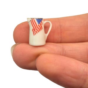 May include: A miniature white pitcher with a handle, featuring a design of the American flag. The flag design includes red and white stripes and a blue field with white stars. The pitcher has a gold rim.