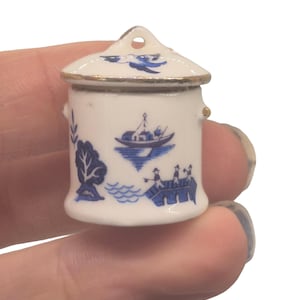 Puede incluir: Pequeño tarro de cerámica blanco y azul con tapa. El tarro presenta un diseño Willow en azul y blanco, que representa un árbol, un barco y un puente. La tapa tiene un pequeño agujero y ribete dorado.