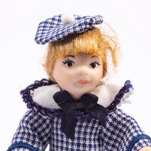 May include: A doll with fair skin, red hair, and brown eyes, wearing a blue and white checkered outfit with a matching hat. The doll has a white collar and a dark blue bow.
