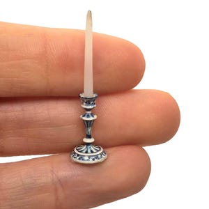 May include: Miniature blue and white ceramic candlestick with a white candle. The candlestick has a detailed design with a round base, a slender stem, and a small candle holder. The candle is white and has a slightly pointed tip. The overall design is delicate and ornate.