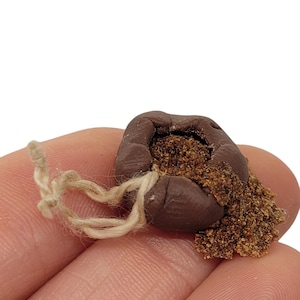 Könnte beinhalten: Ein kleines, braunes, beutelartiges Objekt, gefüllt mit körnigem Material, möglicherweise ein Gewürz oder Kraut. Der Beutel ist mit einer beigen Schnur gebunden. Das Objekt wird in einer Hand vor weißem Hintergrund gehalten.