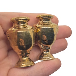 Puede incluir: Dos pequeños jarrones decorativos dorados con forma de urna clásica. Los jarrones tienen una superficie brillante y reflectante y un diseño detallado y texturizado. Se sostienen en una mano sobre un fondo blanco.