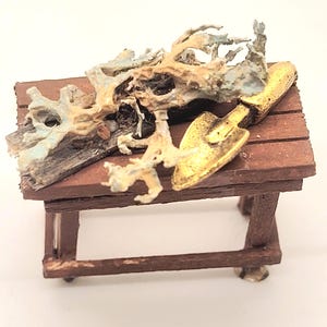 May include: Miniature brown wooden table with a gold-colored shovel and a piece of driftwood. The driftwood is light brown and blue. The table has four legs and is made of wooden planks.