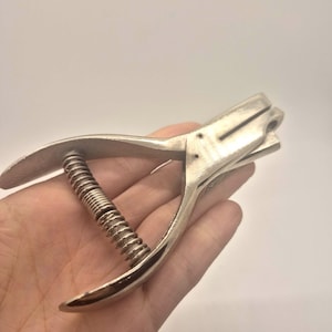 Pode incluir: Uma ferramenta manual de perfuração de cor prateada. A ferramenta possui um mecanismo de mola e alças curvas para agarrar. O perfurador tem uma cabeça retangular com um orifício para perfurar papel.