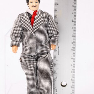 May include: A doll dressed in a gray and white checkered suit with a red tie. The doll is standing next to a ruler that measures 6 inches.