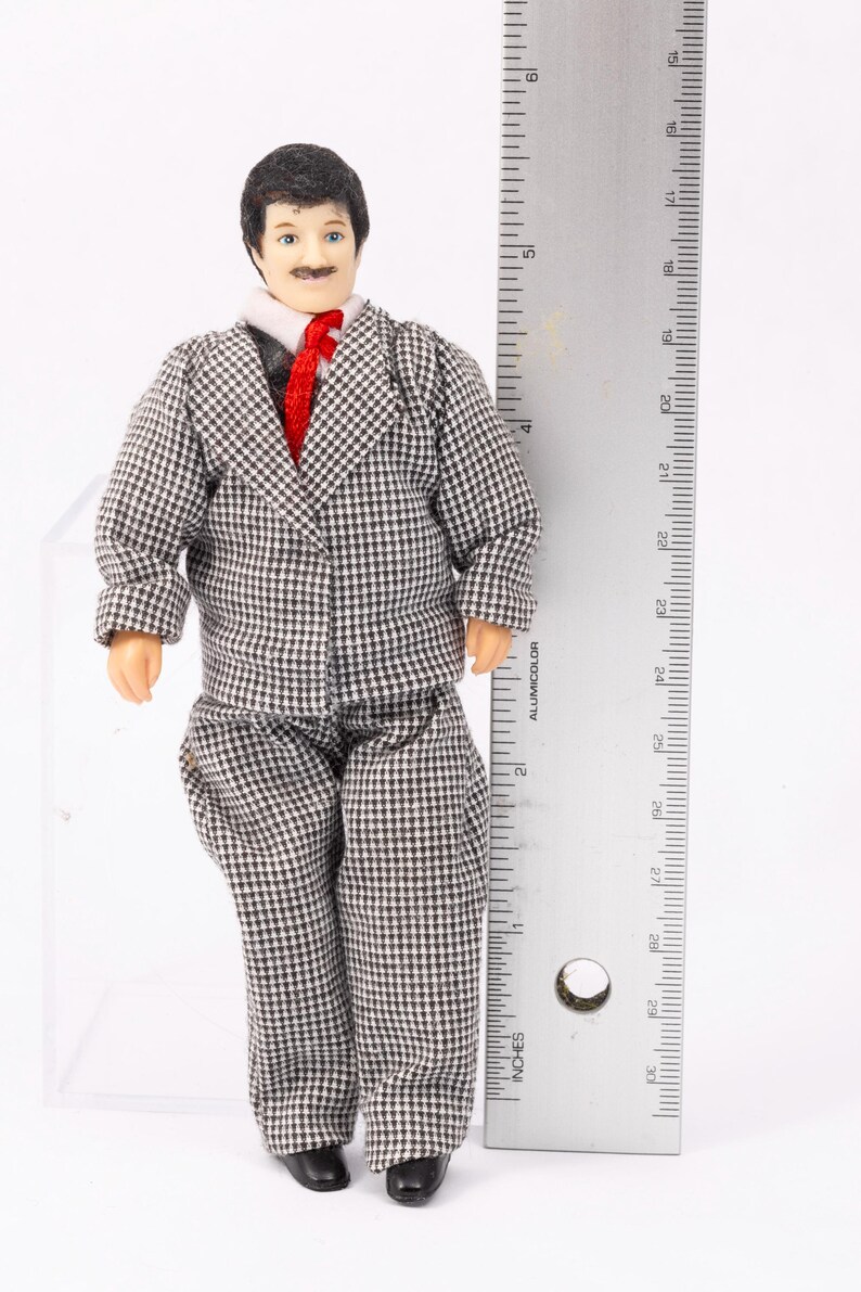 May include: A doll dressed in a gray and white checkered suit with a red tie. The doll is standing next to a ruler that measures 6 inches.