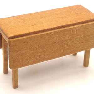 May include: A miniature wooden drop-leaf table with a light brown finish. The table has a rectangular top with rounded corners and four straight legs. The drop-leaf is extended, and the table measures approximately 4 inches long.