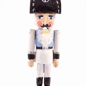 Könnte beinhalten: Holz-Nussknacker-Figur in Form eines Soldaten. Die Figur hat einen schwarzen Hut mit Anker, einen weißen Bart und eine blau-weiße Uniform. Sie steht auf einem kleinen Holzsockel. Die Höhe beträgt ca. 15 cm.