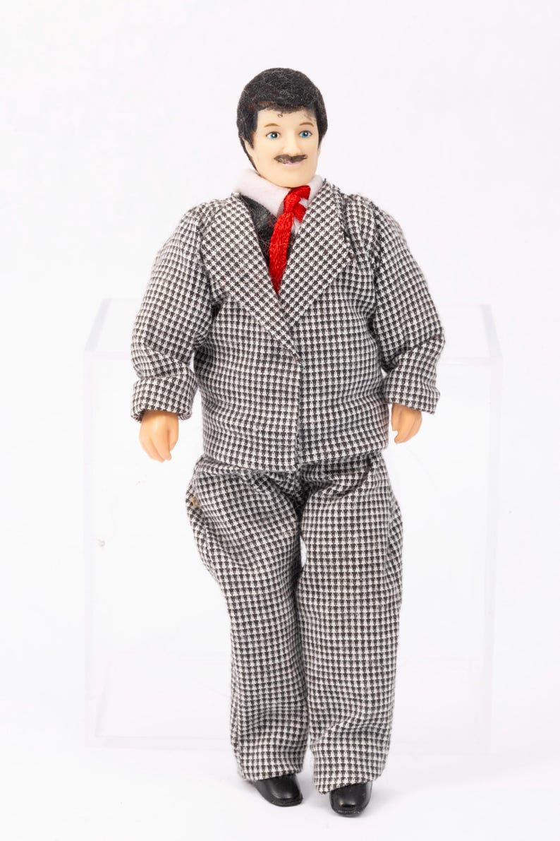 May include: A miniature doll wearing a black and white checkered suit with a red tie. The doll has brown hair and a mustache.