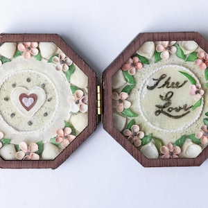 May include: An open, hinged, wooden box with an octagonal shape. The interior features floral and heart designs. One side has the words "Thee Love" in script. The box is decorated with pink flowers and white accents.