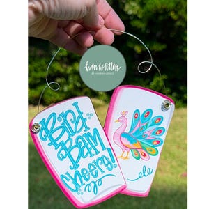 May include: Two white rectangular ornaments with pink edges are held by a wire. One ornament has the words "Bird Bam Cheers!" in turquoise script. The other has a colorful peacock illustration and the word "ele".