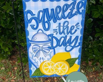 May include: A garden flag with a blue and white gingham pattern and the words "Squeeze the Day" in script. The flag features a lemon-themed design with a jar, lemons, and leaves. A small artist's palette and paintbrush are also visible.