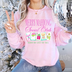 K&ouml;nnte beinhalten: Ein hellrosa Sweatshirt mit dem Text "MERRY MAHJONG Social Club" in rosa Schrift. Darunter befinden sich vier illustrierte Mahjong-Kacheln und der Satz "WHERE WE DECK THE TILES". Die Person tr&auml;gt eine blaue Jeans.