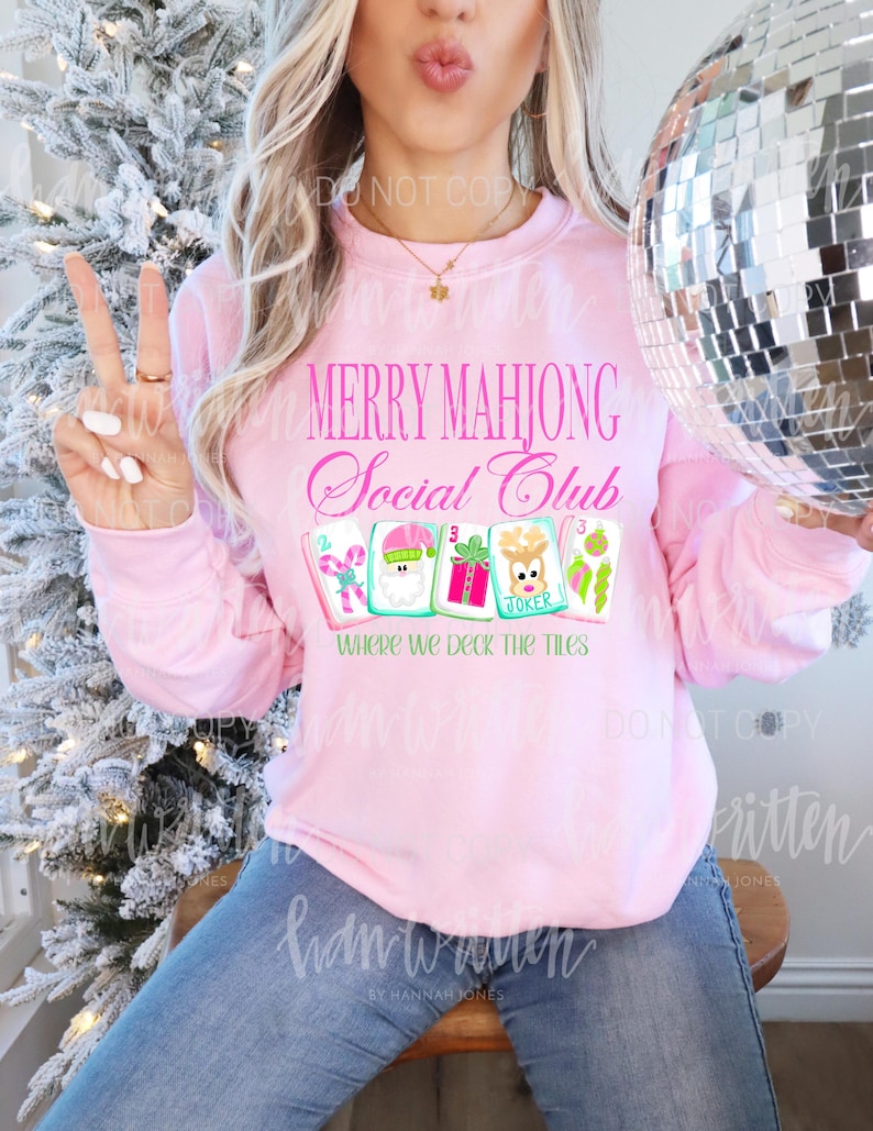 K&ouml;nnte beinhalten: Ein hellrosa Sweatshirt mit dem Text "MERRY MAHJONG Social Club" in rosa Schrift. Darunter befinden sich vier illustrierte Mahjong-Kacheln und der Satz "WHERE WE DECK THE TILES". Die Person tr&auml;gt eine blaue Jeans.