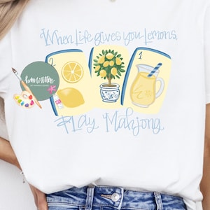 May include: White t-shirt with a colorful graphic design featuring lemons, a lemon tree, and a pitcher of lemonade. The text reads "When life gives you lemons, play Mahjong."