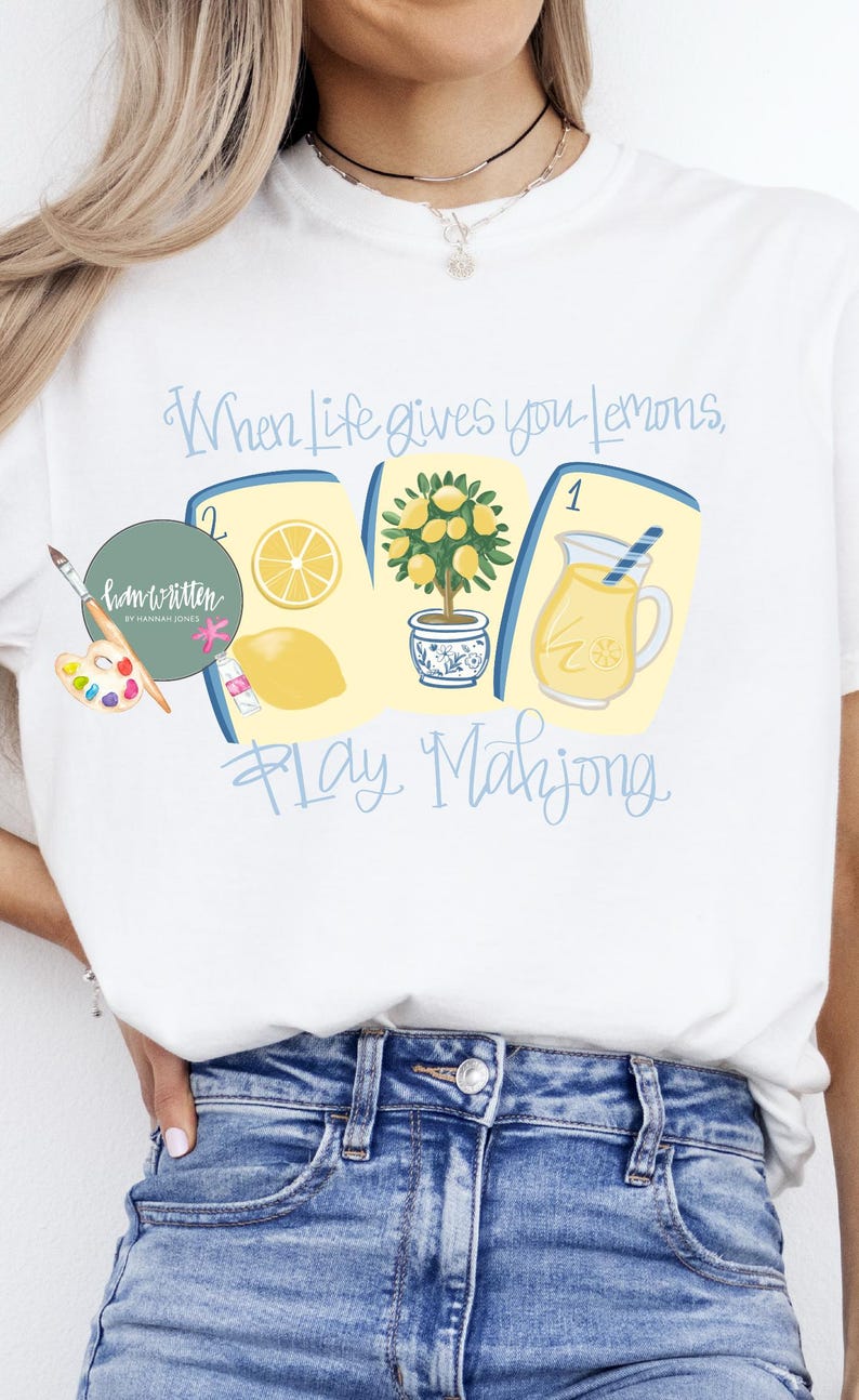 May include: White t-shirt with a colorful graphic design featuring lemons, a lemon tree, and a pitcher of lemonade. The text reads "When life gives you lemons, play Mahjong."