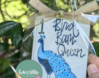 May include: A decorative sign featuring a blue peacock with the text "Bird Bam Queen." The sign is wrapped in clear cellophane and tied with a gold ribbon. The sign also includes a watercolor paint palette and the text "hand written by Hannah Jones."