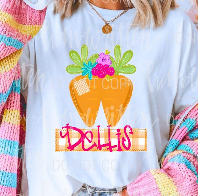 Puede incluir: Camiseta blanca con un dise&ntilde;o de zanahoria de dibujos animados. La zanahoria tiene hojas verdes, flores rosas y azules y un parche a cuadros. La palabra "Dellis" est&aacute; escrita en rosa sobre una pancarta a cuadros. Ropa casual.