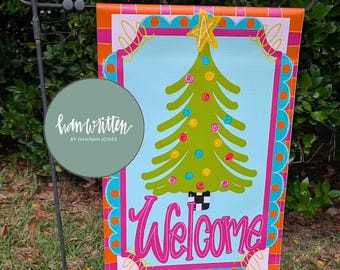May include: A rectangular garden flag featuring a light blue center with a green Christmas tree decorated with colorful ornaments and a yellow star. The word Welcome is written in pink script at the bottom, framed by a bright orange and teal border.