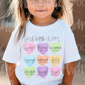 May include: A white t-shirt with the text "God says I am..." printed on the front. The shirt features nine colorful hearts with words and bible verses inside each heart. The words include "beautiful", "strong", "courageous", "treasured", "redeemed", "bold", "unique", "loved", and "loyal".