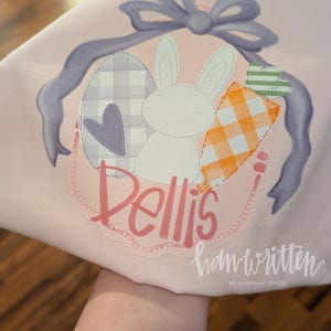 May include: A pink Easter-themed t-shirt with a purple bow, a white bunny, a checkered egg, a heart, and a carrot. The shirt has the name "Bellis" written in pink cursive and the text "handwritten by Hannah Jones" in black cursive.