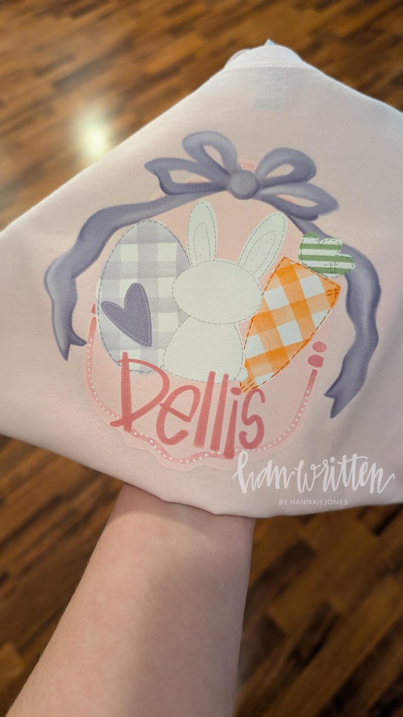 May include: A pink Easter-themed t-shirt with a purple bow, a white bunny, a checkered egg, a heart, and a carrot. The shirt has the name "Bellis" written in pink cursive and the text "handwritten by Hannah Jones" in black cursive.