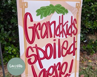 May include: A rectangular garden flag featuring a light tan background with white polka dots and a gold border. The text "Grandkids Spoiled Here!" is written in a bold red script font, accented by green leafy illustrations at the top and bottom.