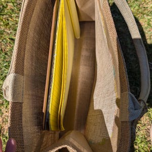 May include: A tan burlap tote bag with a rectangular shape and two handles. The bag is open, revealing yellow legal pads and notebooks inside. The bag is sitting on green grass.