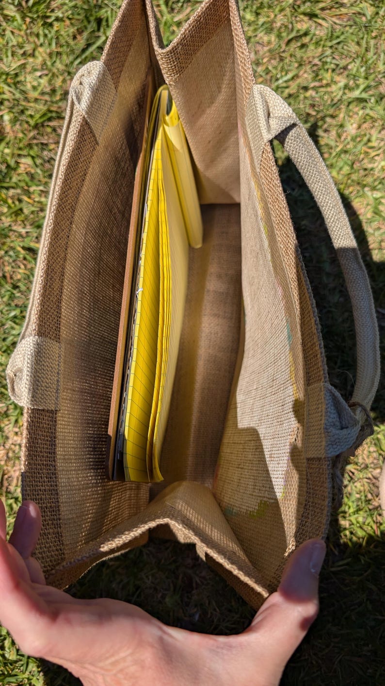 May include: A tan burlap tote bag with a rectangular shape and two handles. The bag is open, revealing yellow legal pads and notebooks inside. The bag is sitting on green grass.