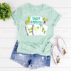 May include: A pale green t-shirt featuring the words "Cinco de Mahjong" and mahjong tile illustrations, a margarita, and cacti. The shirt is styled with denim shorts and white trainers.