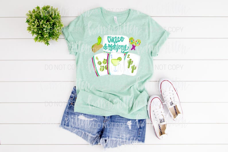 May include: A pale green t-shirt featuring the words "Cinco de Mahjong" and mahjong tile illustrations, a margarita, and cacti. The shirt is styled with denim shorts and white trainers.