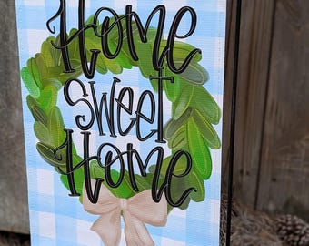 May include: A garden flag with a light blue and white checkered background. The flag features the words "Home Sweet Home" in black script, surrounded by a green wreath and a beige bow. The flag is displayed on a black metal stand.