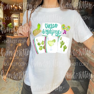 May include: White t-shirt with a colourful graphic design featuring the text "Cinco & Mahjong" and illustrations of a taco, flowers, and playing cards. The shirt is worn by a person.