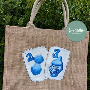 May include: A tan burlap tote bag with cream-coloured handles. The front of the bag features two white playing cards with blue painted designs and numbers. The text "ham written by Hannah Jones" is on a green circle.