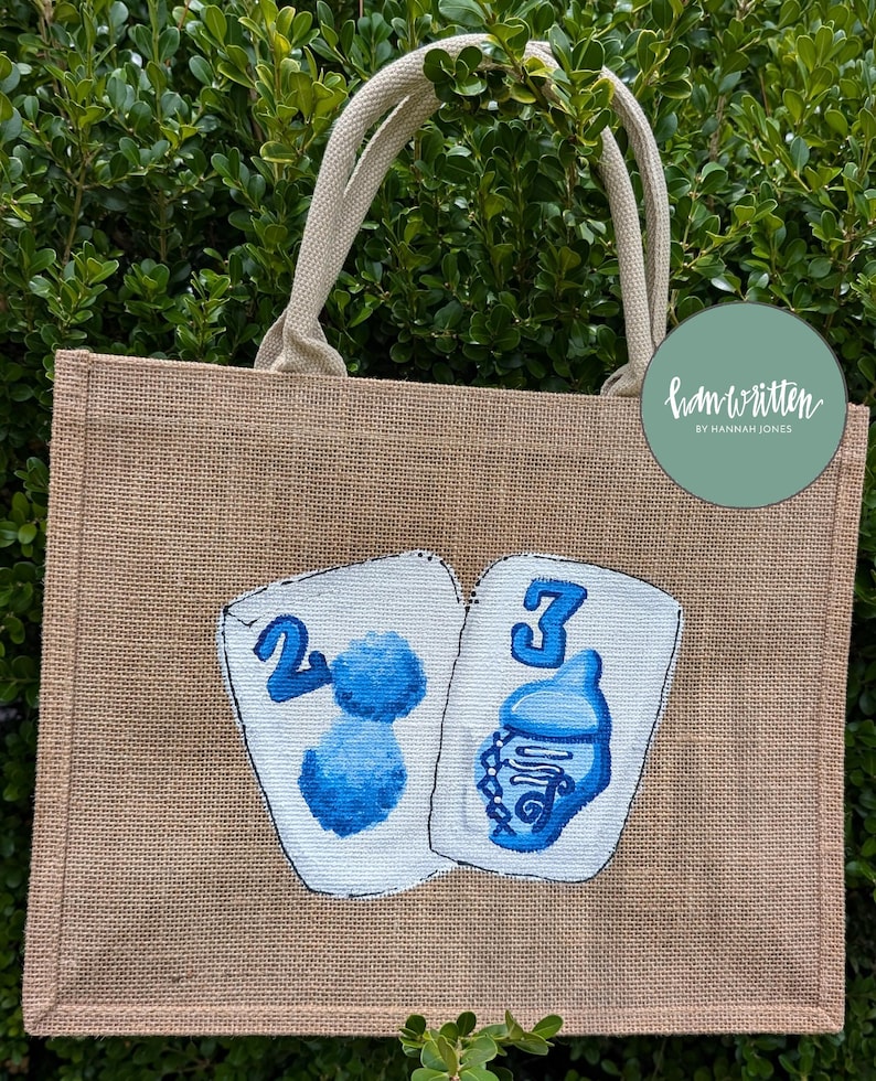 May include: A tan burlap tote bag with cream-coloured handles. The front of the bag features two white playing cards with blue painted designs and numbers. The text "ham written by Hannah Jones" is on a green circle.