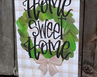 May include: A garden flag with a tan and white checkered background. The flag features a green wreath with the words "Home Sweet Home" in black script. A light pink bow is at the bottom of the wreath.