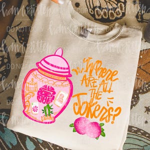 May include: A beige sweatshirt featuring a pink and orange illustration of a decorative jar with the text "Where Are All The Jokers?" in orange. The design includes pink peaches and green leaves.