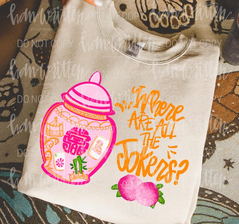 May include: A beige sweatshirt featuring a pink and orange illustration of a decorative jar with the text "Where Are All The Jokers?" in orange. The design includes pink peaches and green leaves.