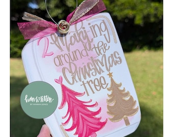 May include: A white Christmas ornament with the text "2 Walking around the Christmas tree". The ornament features a pink Christmas tree and a gold Christmas tree. It has a pink and gold ribbon and a wire loop for hanging.