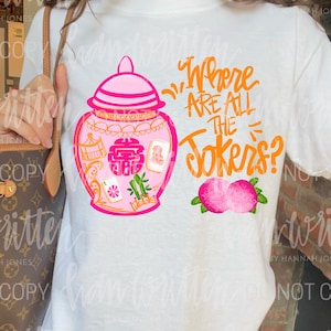 May include: White t-shirt featuring a pink and orange graphic. The design includes a pink jar with a lid, a double happiness symbol, and the text "Where Are All The Jokers?" in orange, along with two pink peaches.